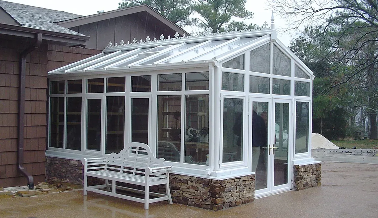 A white sunroom with glass walls and a stone base, attached to a brown house. A white bench sits outside.