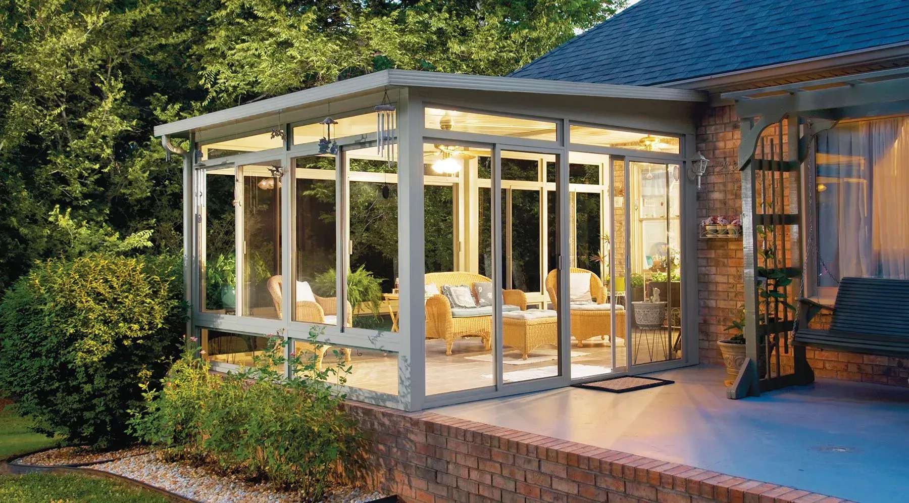Sunroom with glass walls and roof, attached to a brick home, with seating inside.