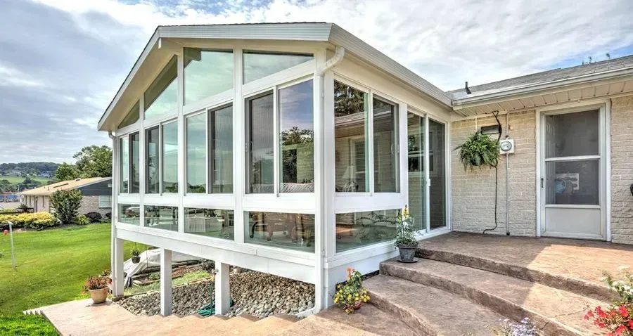 White sunroom with large windows, built on a raised foundation, beside a home.
