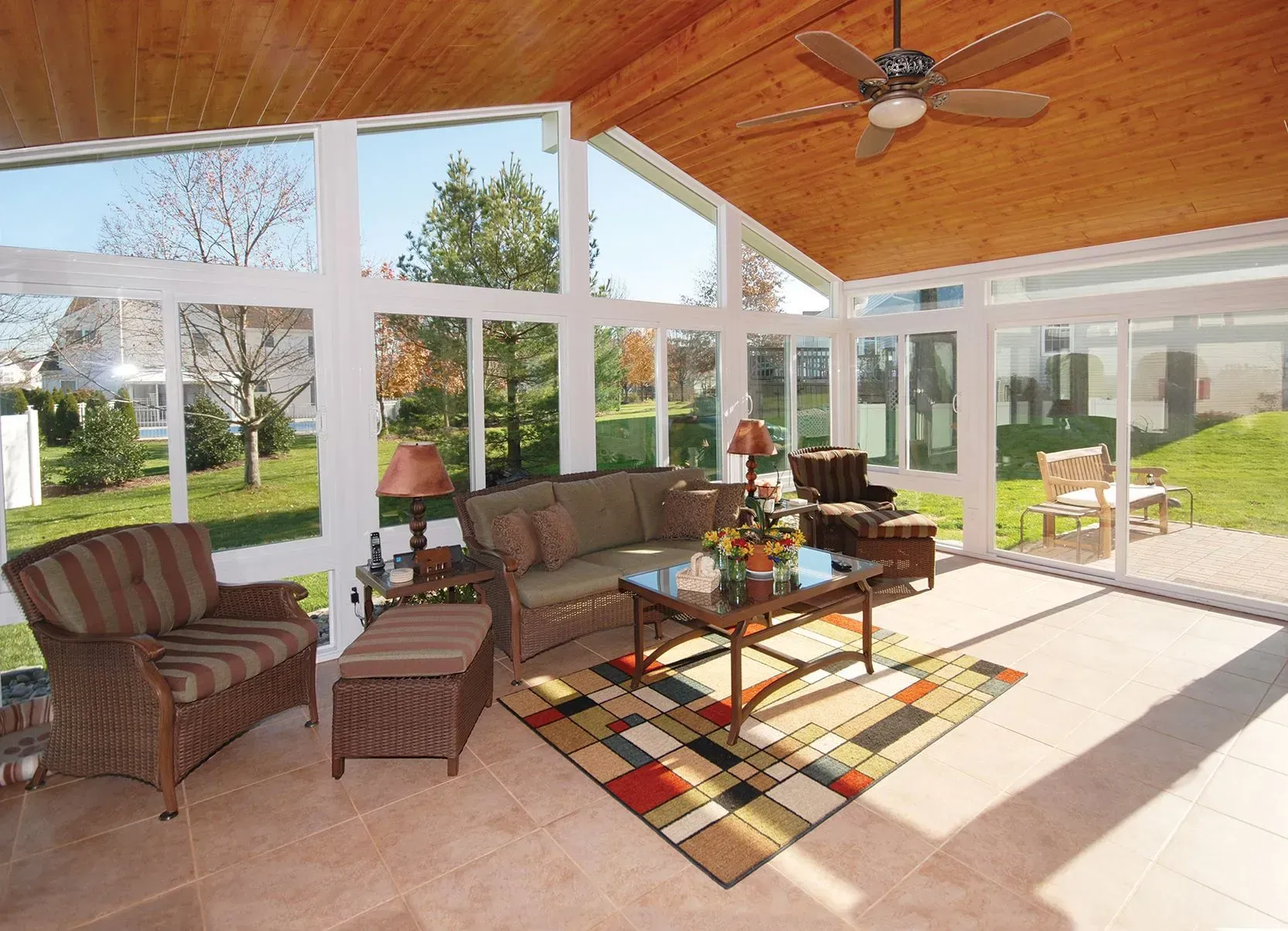 Sunroom with glass walls, wood ceiling, and wicker furniture. Overlooking a green yard.