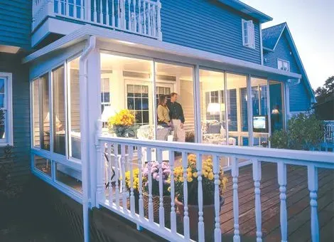 A screened porch attached to a blue house with a couple inside, viewed from a wooden deck with flowers.