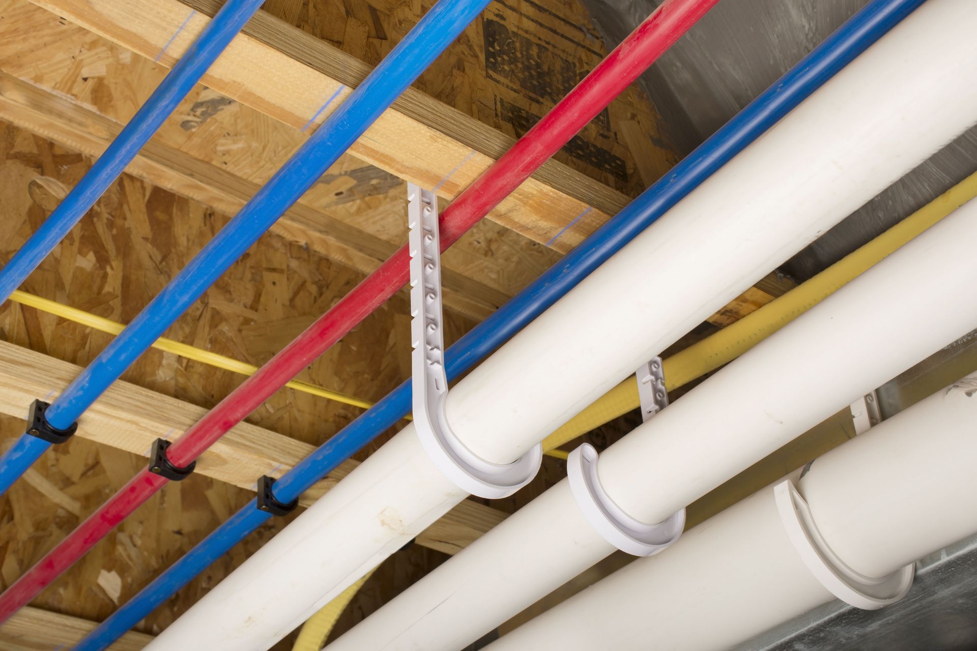 Pipes secured to a wood ceiling, blue and red PEX water lines, white drain pipes, and yellow electrical conduit.