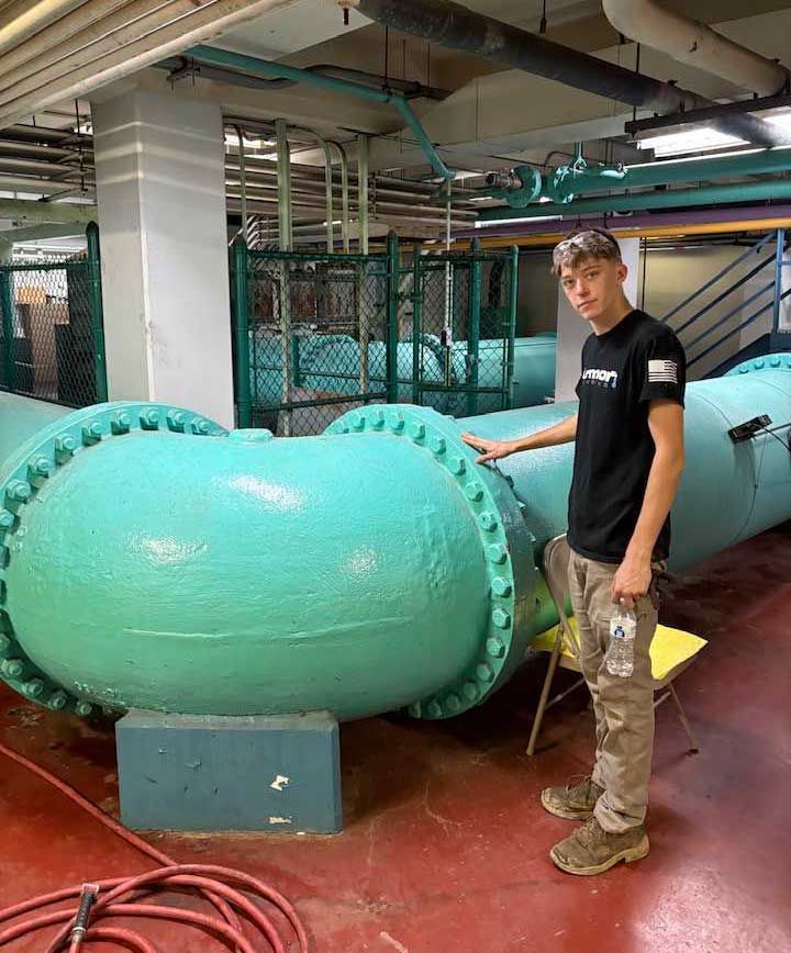 Young man in black shirt and work pants stands near large teal water pipes.