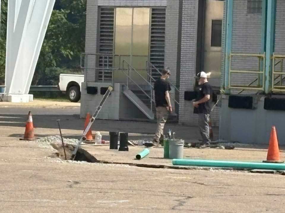 Two workers near a building with a cracked pavement and construction materials; orange cones mark the area.