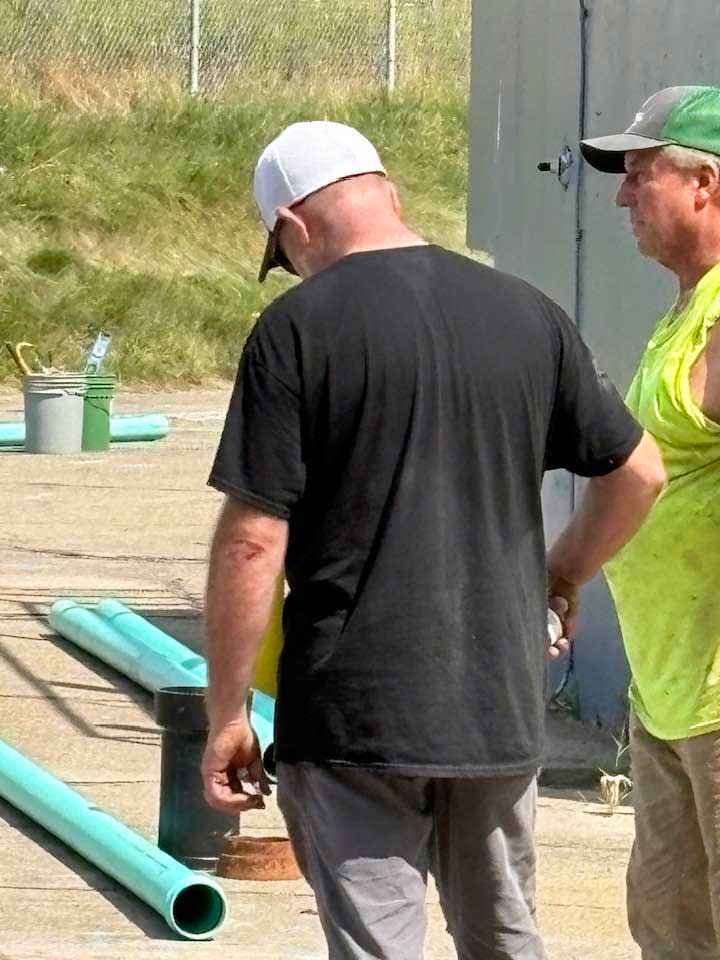 Two men outside: one wearing a white cap and black shirt, the other in a green vest, looking at something.