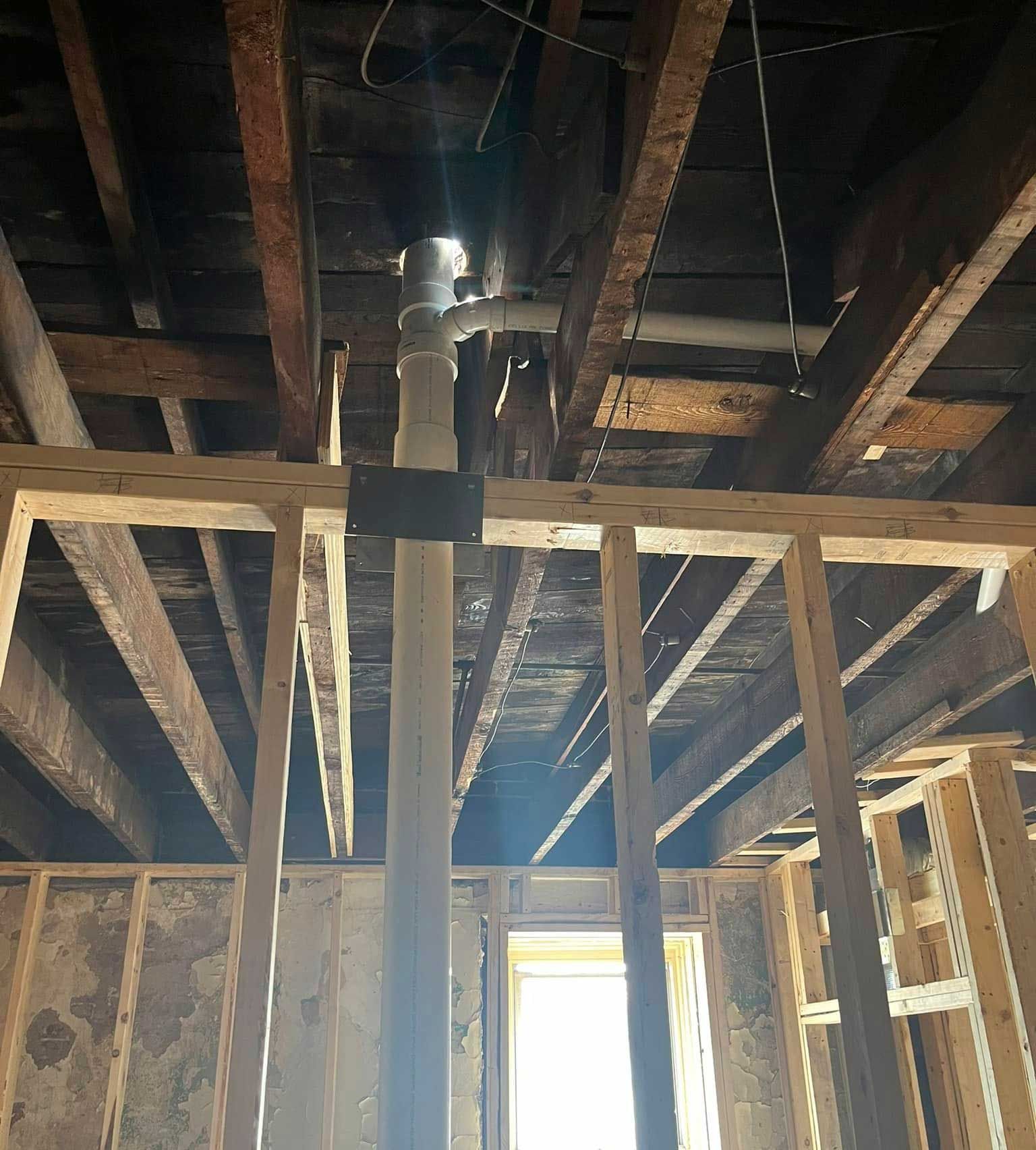 Interior view of plumbing pipes against wooden framing. Bright natural light shines through an opening.