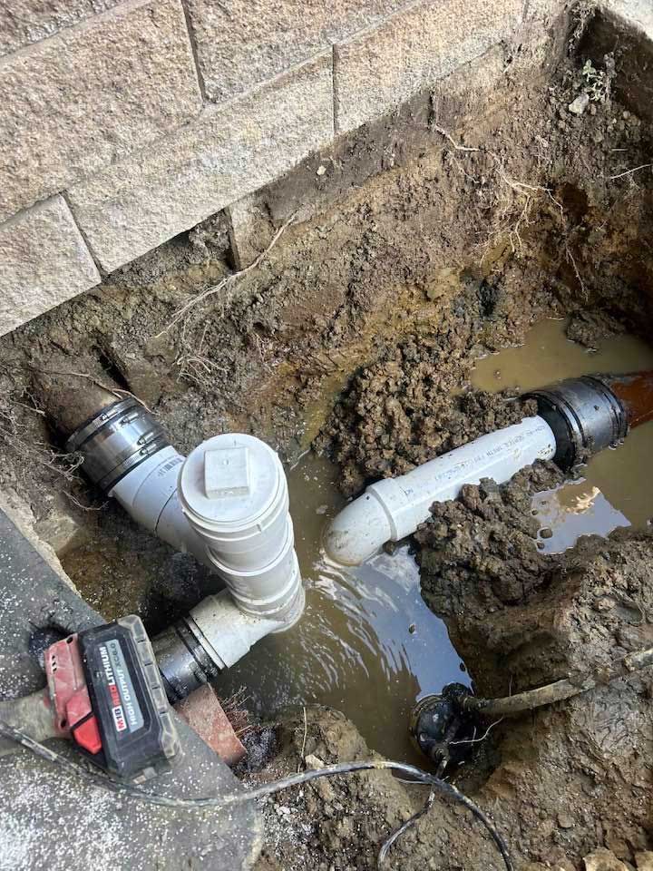 Excavated plumbing pipes in dirt, near a brick wall. White and black pipes with water pooling around them.