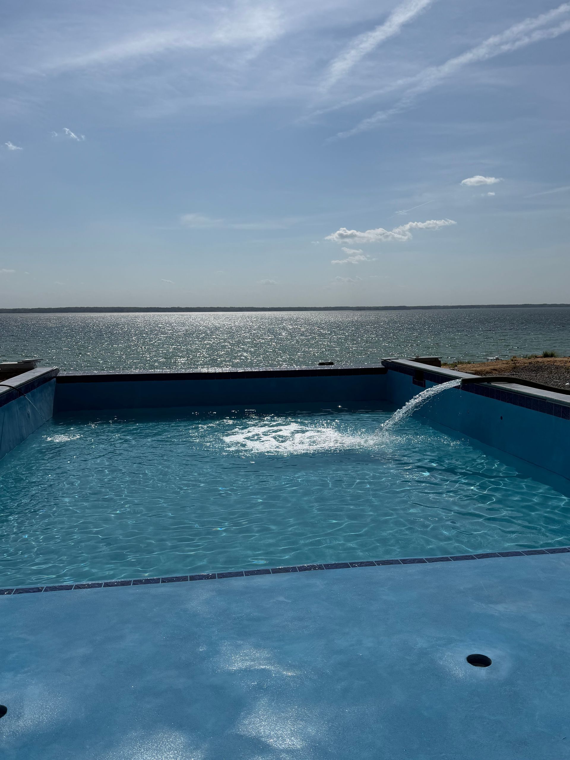 A bright blue rectangular pool sits overlooking a sparkling lake under a clear, sunny sky, with water pouring in.
