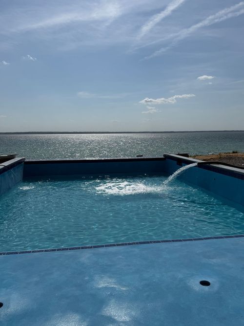 A bright blue rectangular pool sits overlooking a sparkling lake under a clear, sunny sky, with water pouring in.