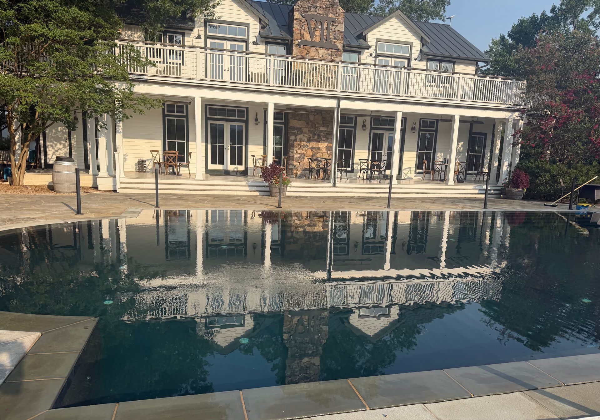 Luxury house reflected in a calm backyard pool with a stone patio and trees nearby