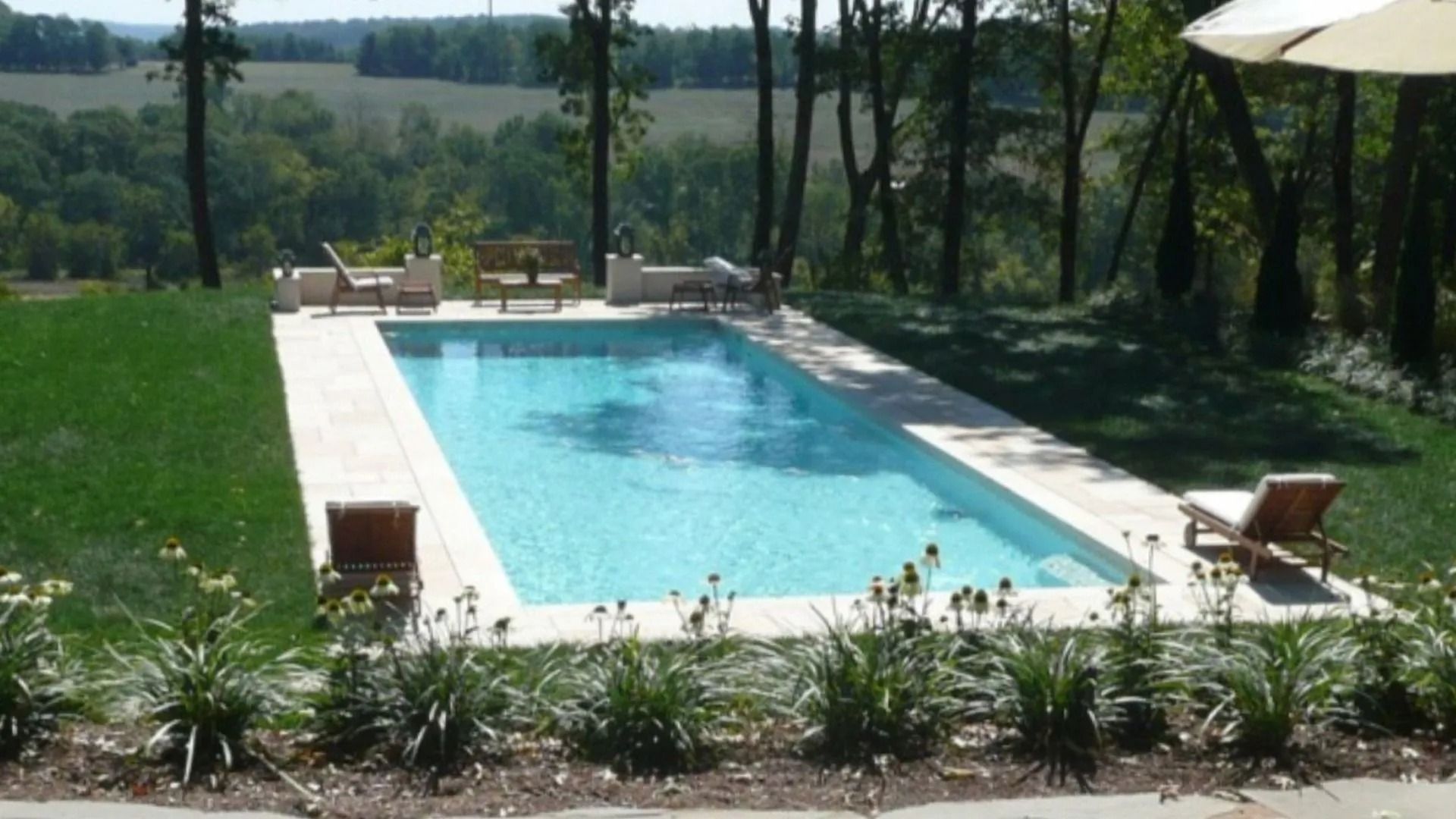 Rectangular pool with light blue water surrounded by beige tiles and green lawn, trees in the background.