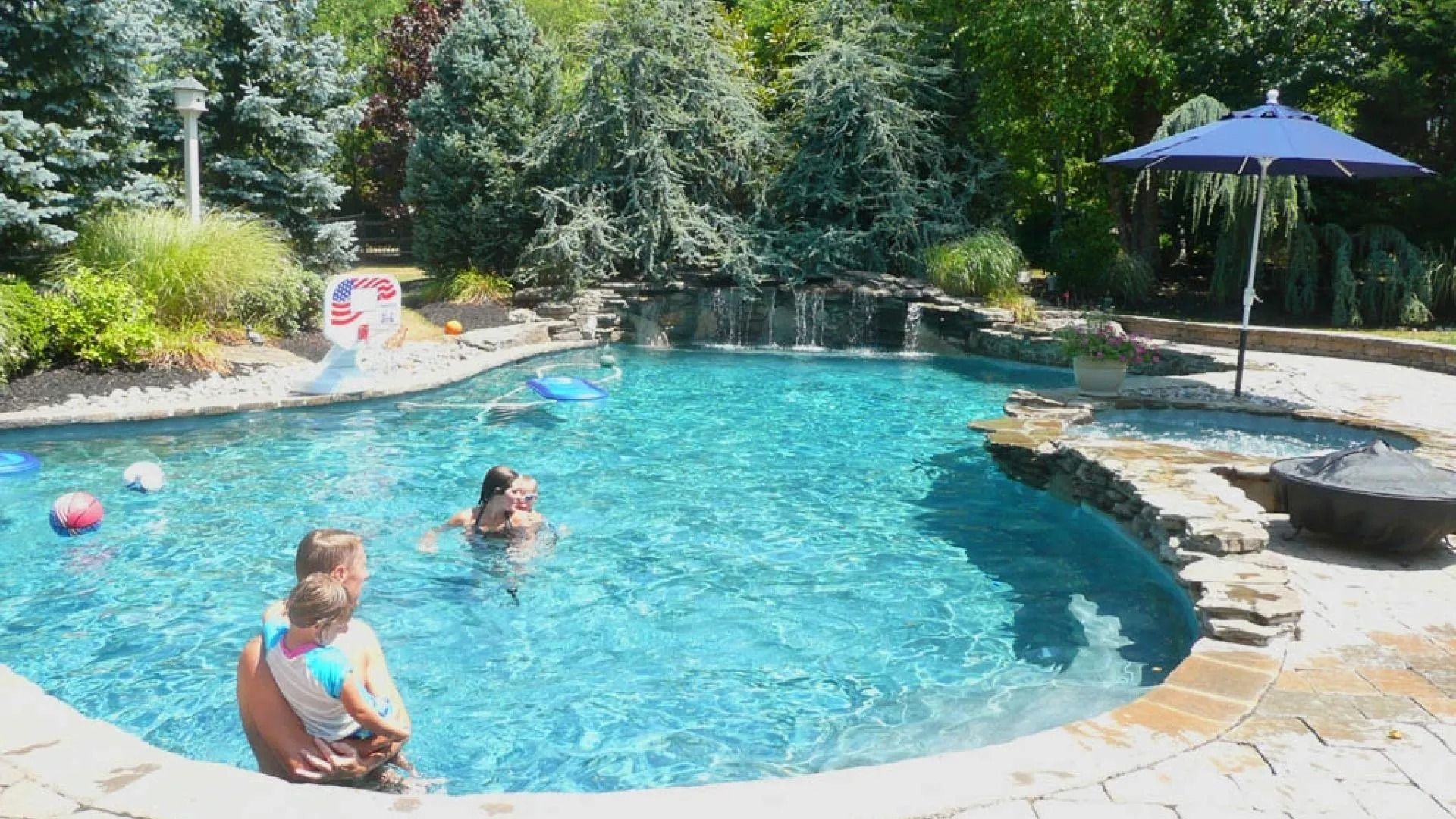Swimming pool with people. Bright blue water, waterfall, umbrella, and a child in arms on the edge. Sunny outdoor setting.