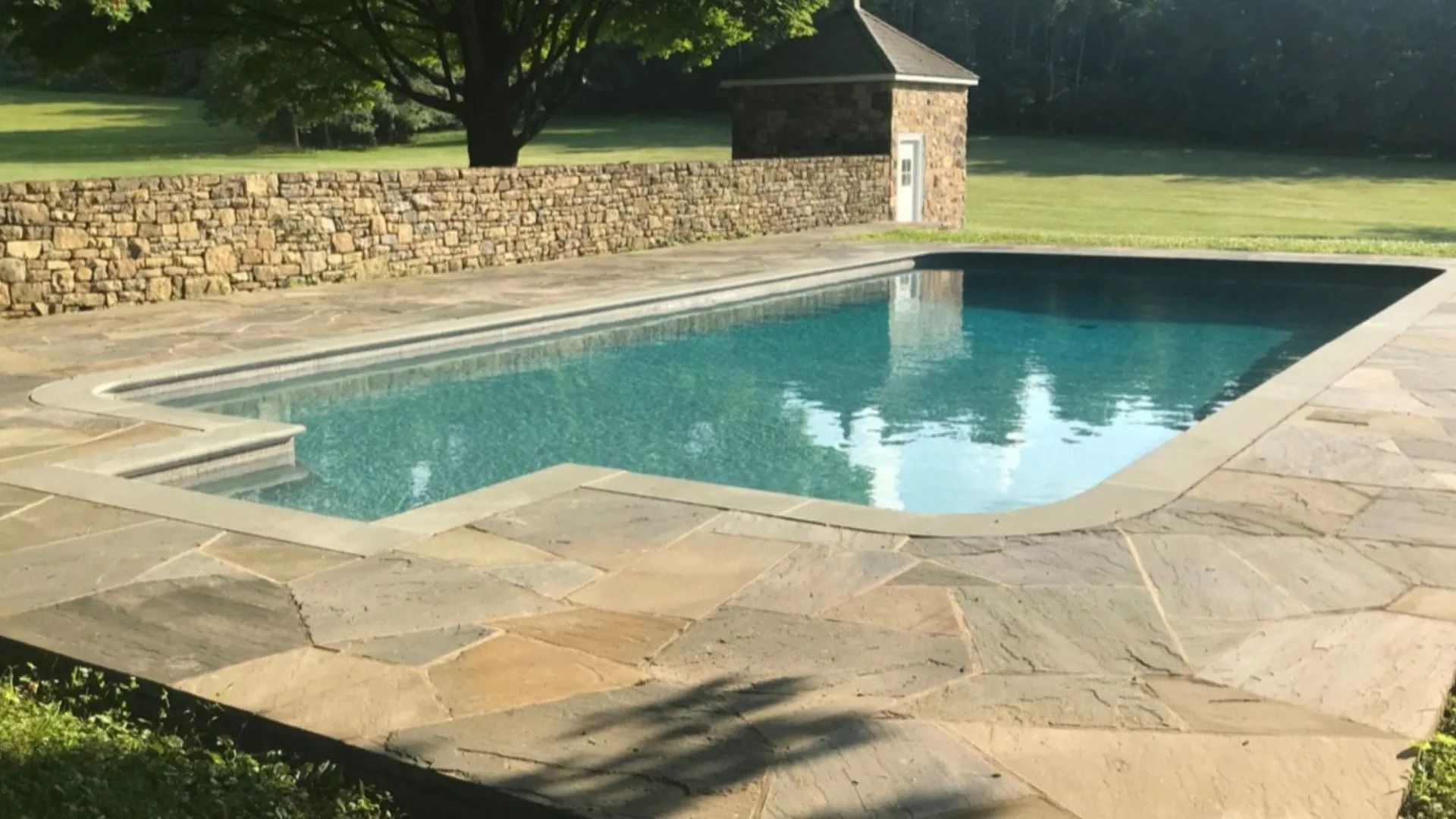 Swimming pool with stone patio, stone wall, and small building in a grassy yard.