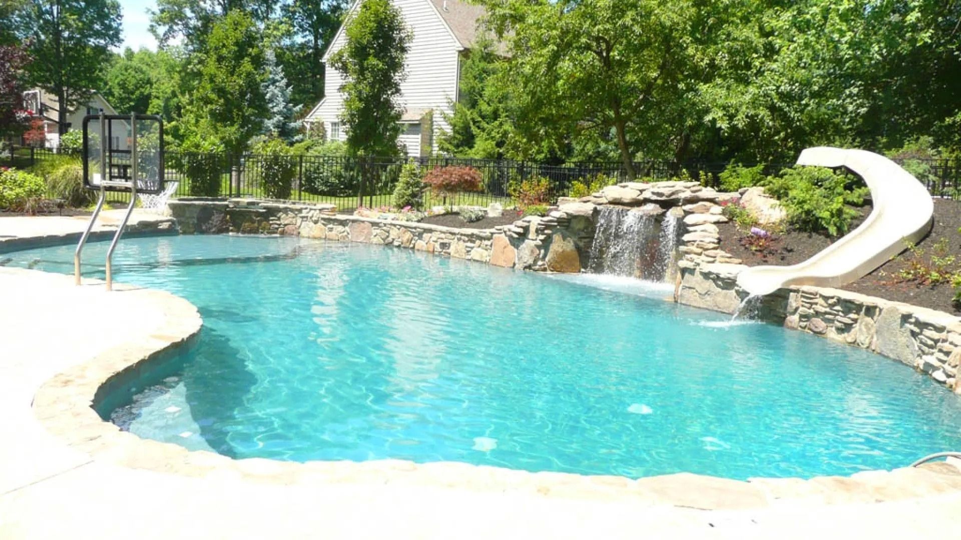 Swimming pool with rock waterfall and slide, surrounded by greenery.