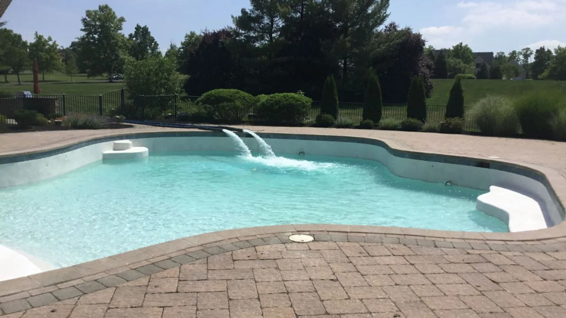 Swimming pool with two waterfalls, surrounded by a brick patio, with trees and grass in the background.