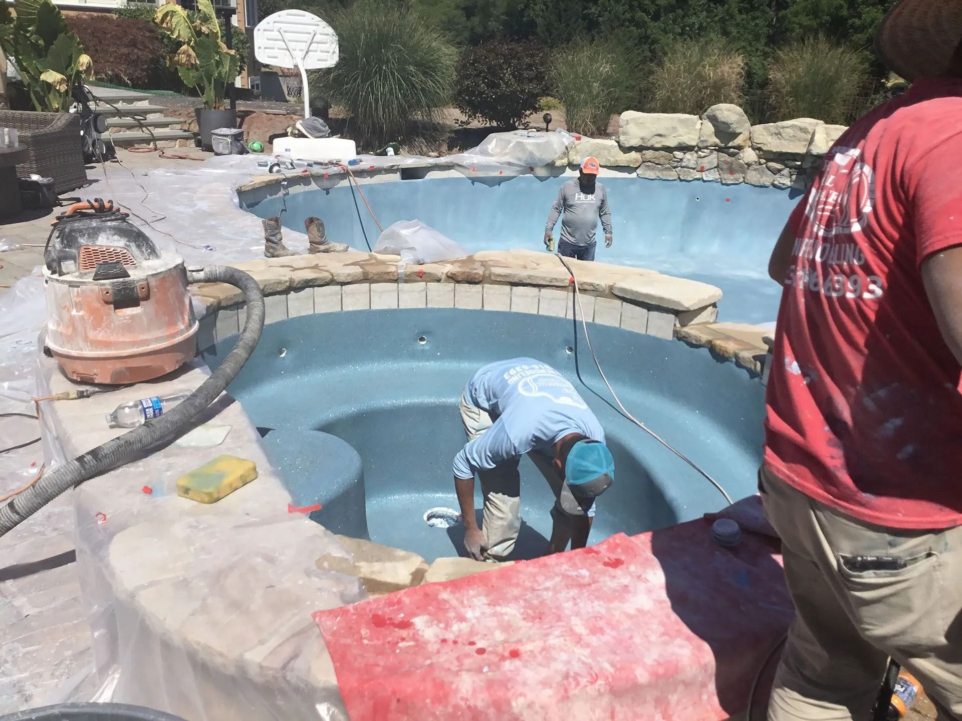 Workers resurfacing a blue-tiled pool with equipment. One person is in the pool; others on the perimeter.