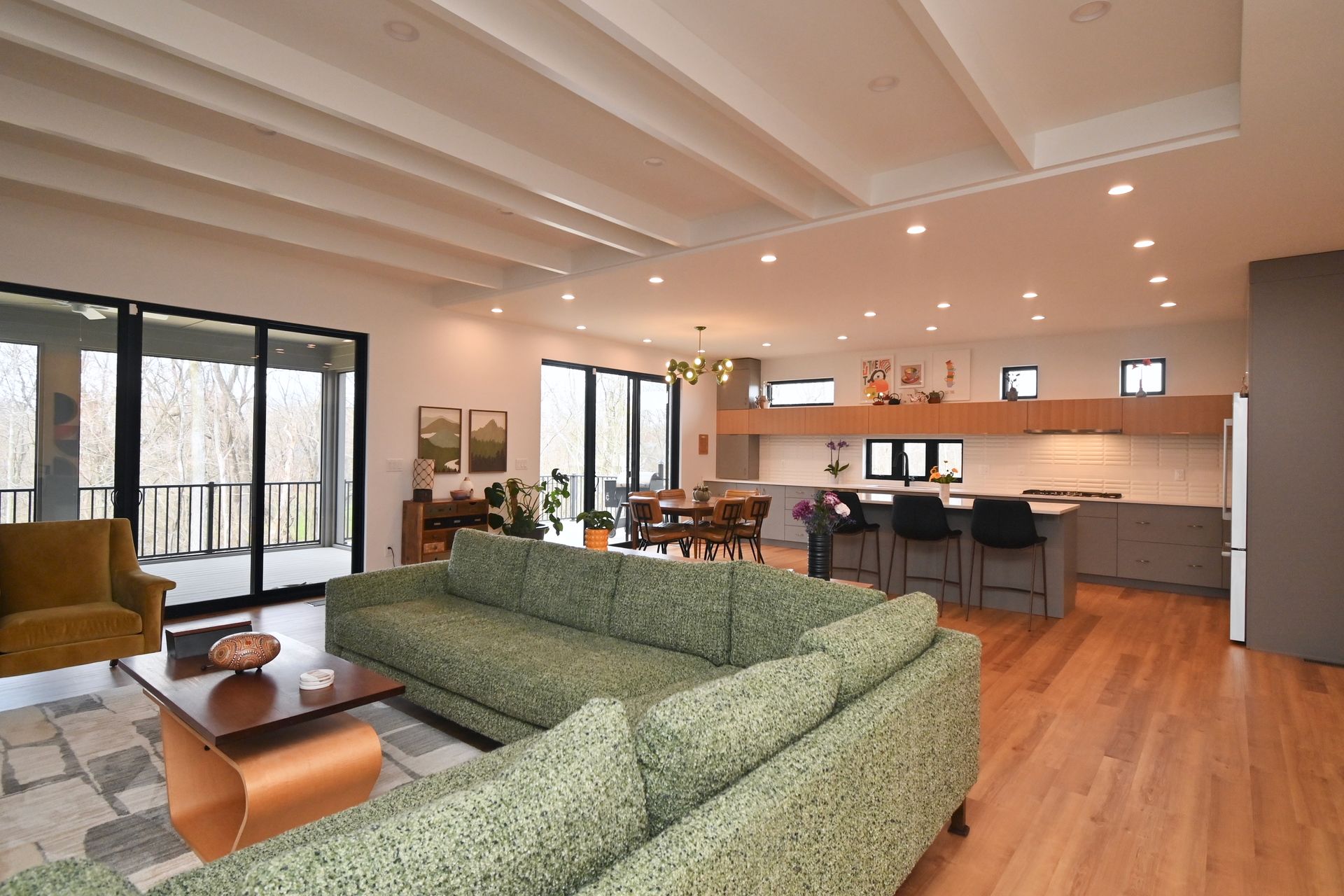 Spacious, modern living room with green sofa, wooden floors, and open kitchen. Large windows overlook a wooded area.