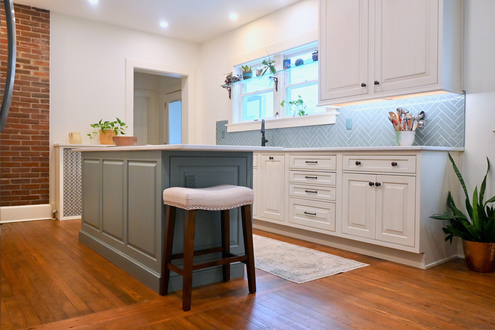 Kitchen with gray island and white cabinets, hardwood floors, brick wall.