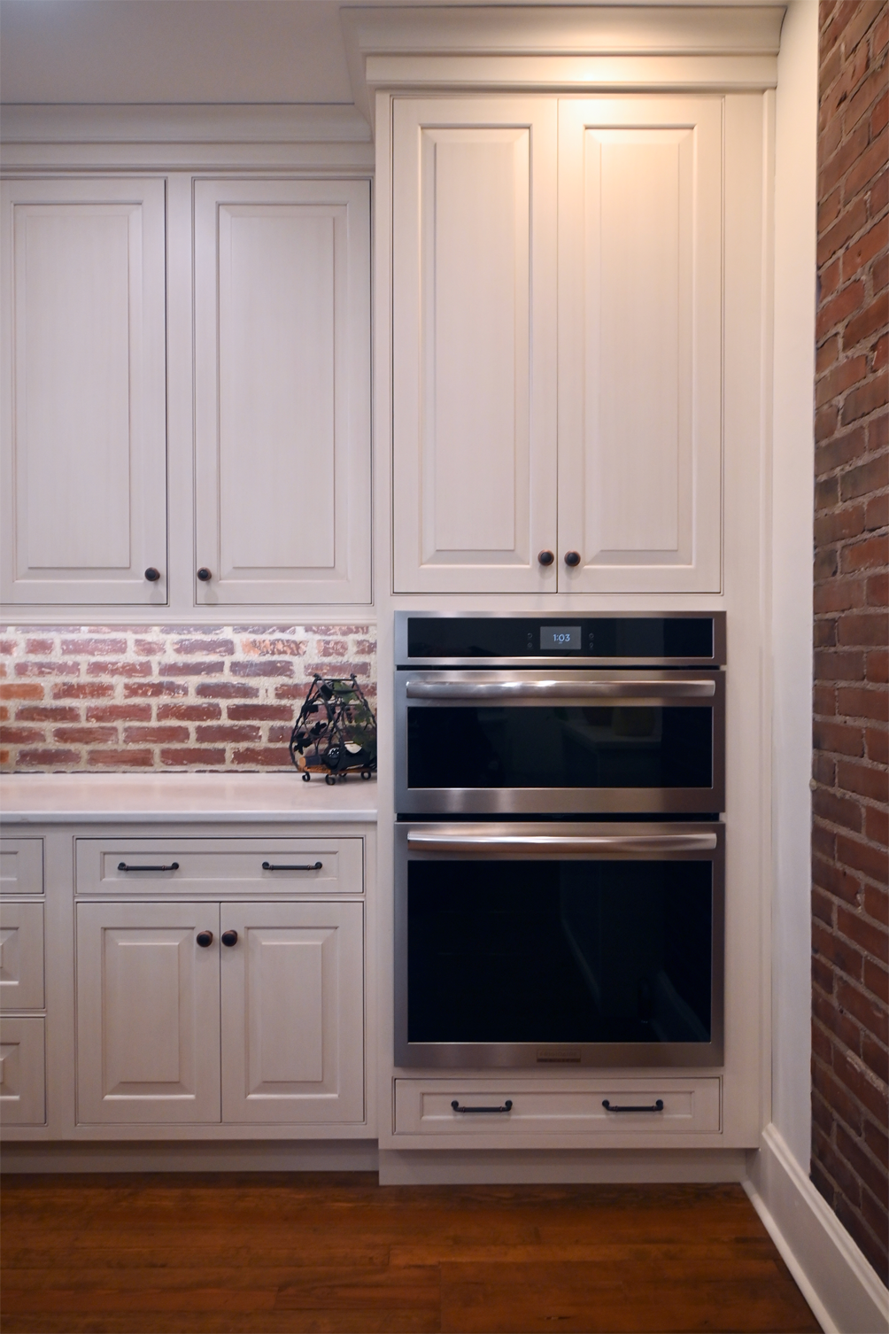 Kitchen cabinet with built-in oven and brick backsplash. White cabinets, stainless steel oven, wood floor.