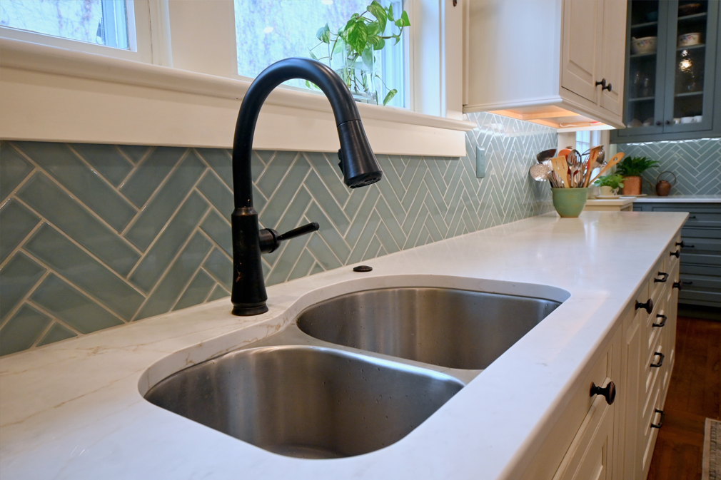 Kitchen with white countertops, a double stainless steel sink, black faucet, and blue herringbone tile backsplash.