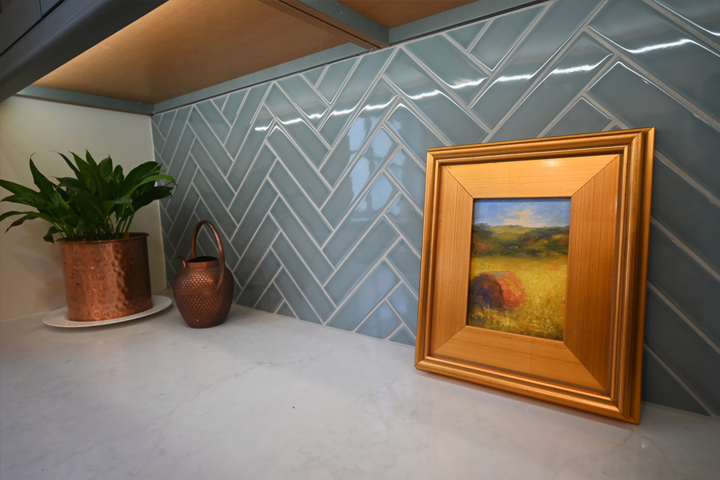 A framed landscape painting rests on a white countertop with a copper watering can, plant, and blue herringbone tile backsplash.