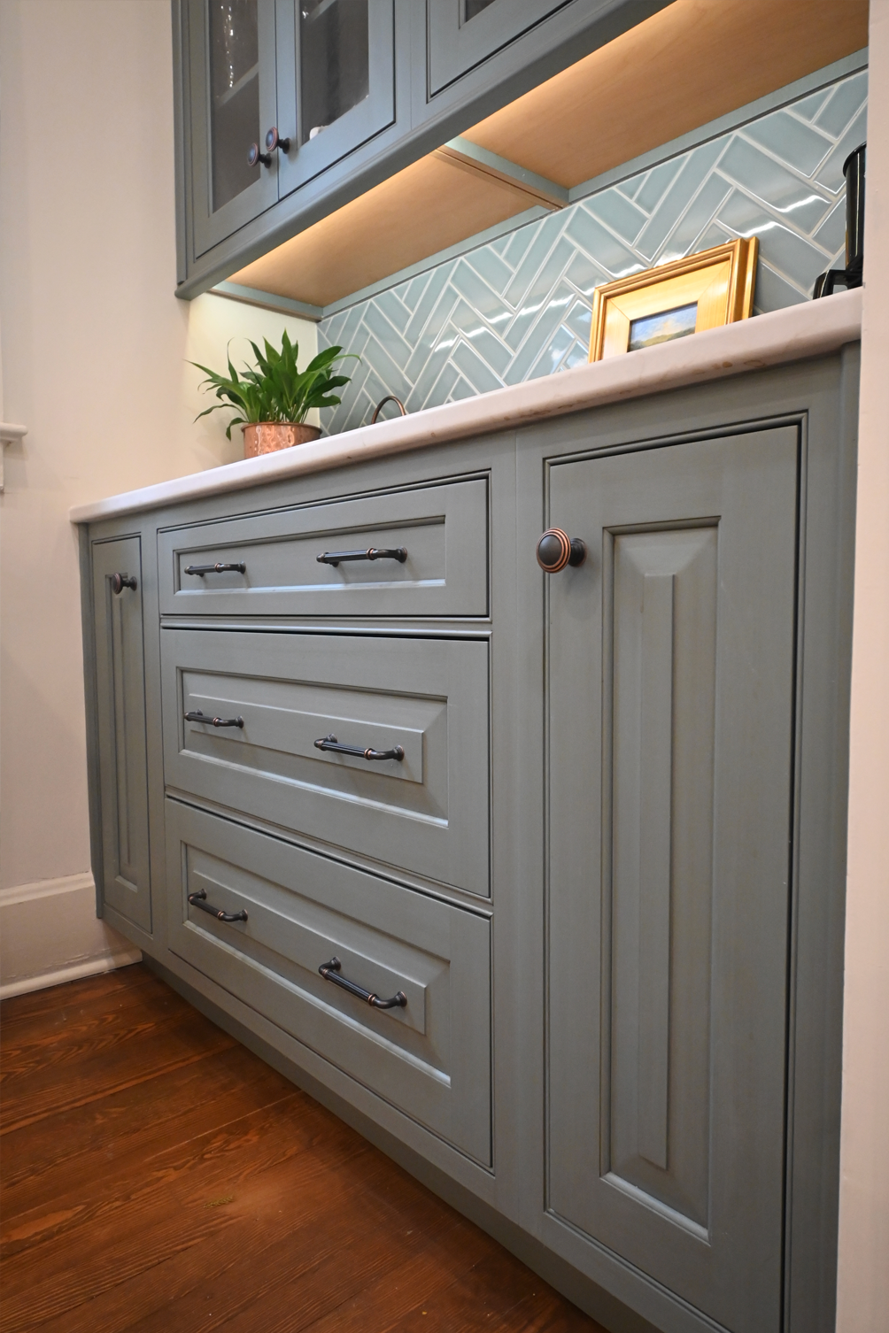 Blue cabinets with drawers, white countertop, backsplash, wooden floor, and a plant.