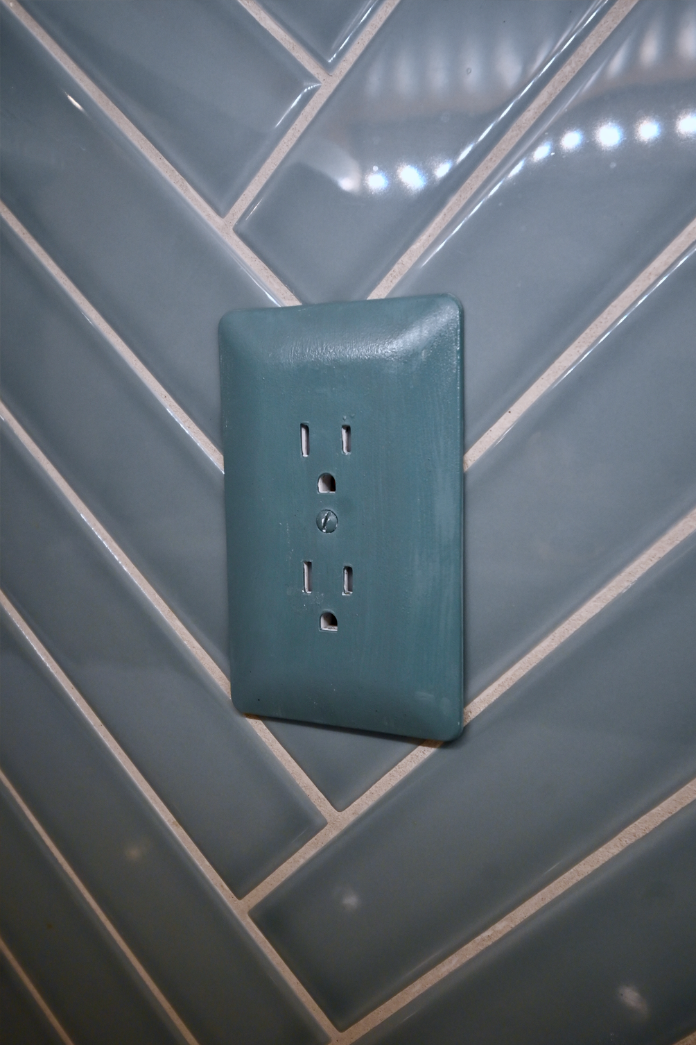 Teal electrical outlet on blue herringbone tile wall.