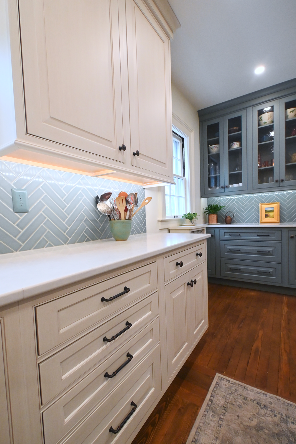 Cream and blue kitchen cabinetry with white countertops, herringbone backsplash, and wood floors.