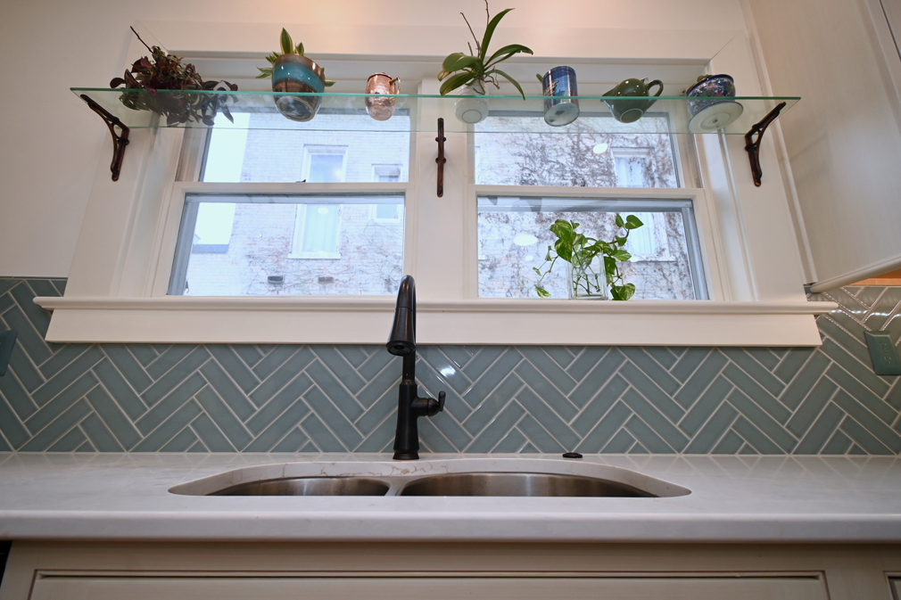 Kitchen sink with a blue herringbone tile backsplash, window with plants on a shelf.