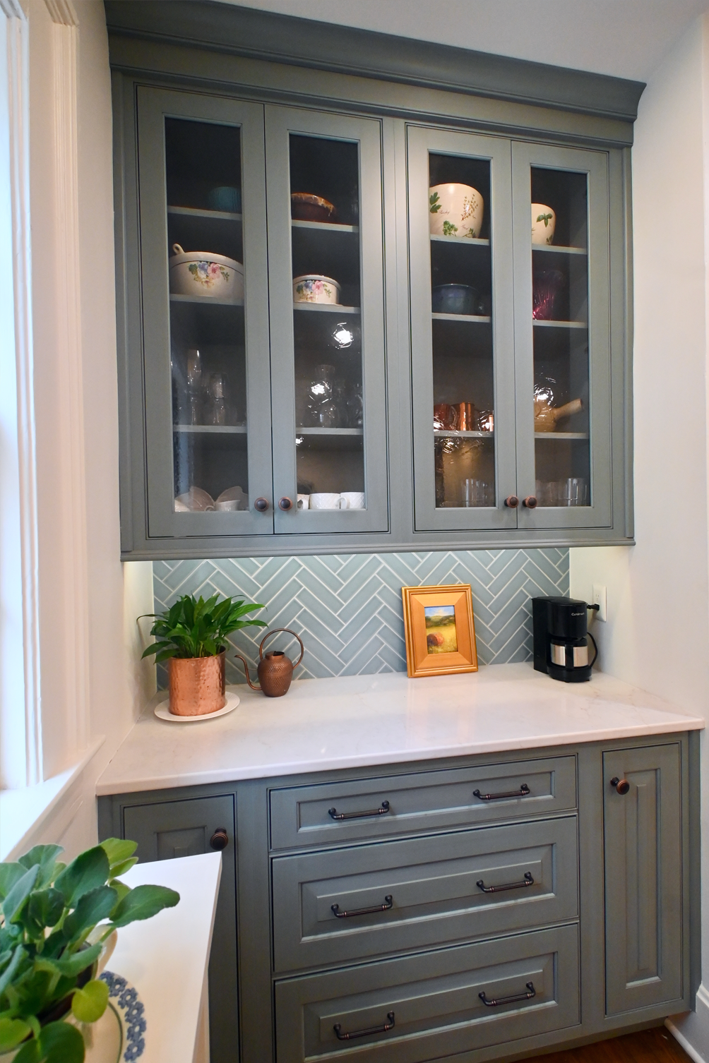 Gray kitchen cabinets with glass doors, white countertop, and herringbone tile backsplash.
