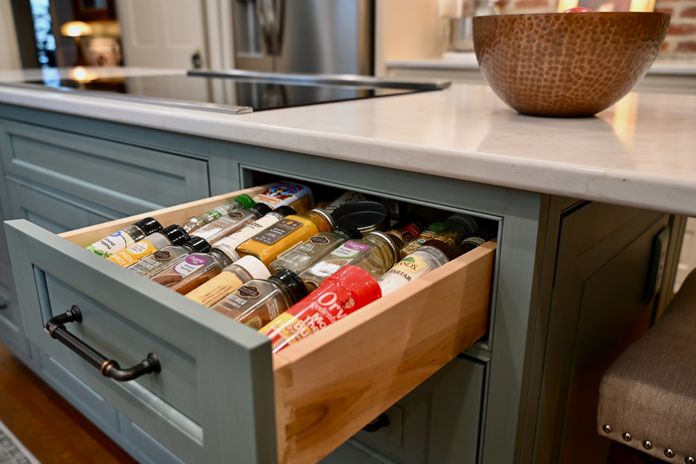 Open kitchen drawer filled with spice jars on a blue island, with a countertop and bowl visible.
