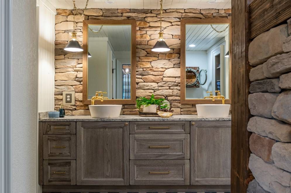A bathroom with two sinks , two mirrors and a stone wall.