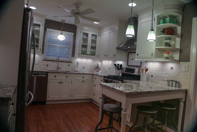 A kitchen with white cabinets , granite counter tops , stools and a ceiling fan.