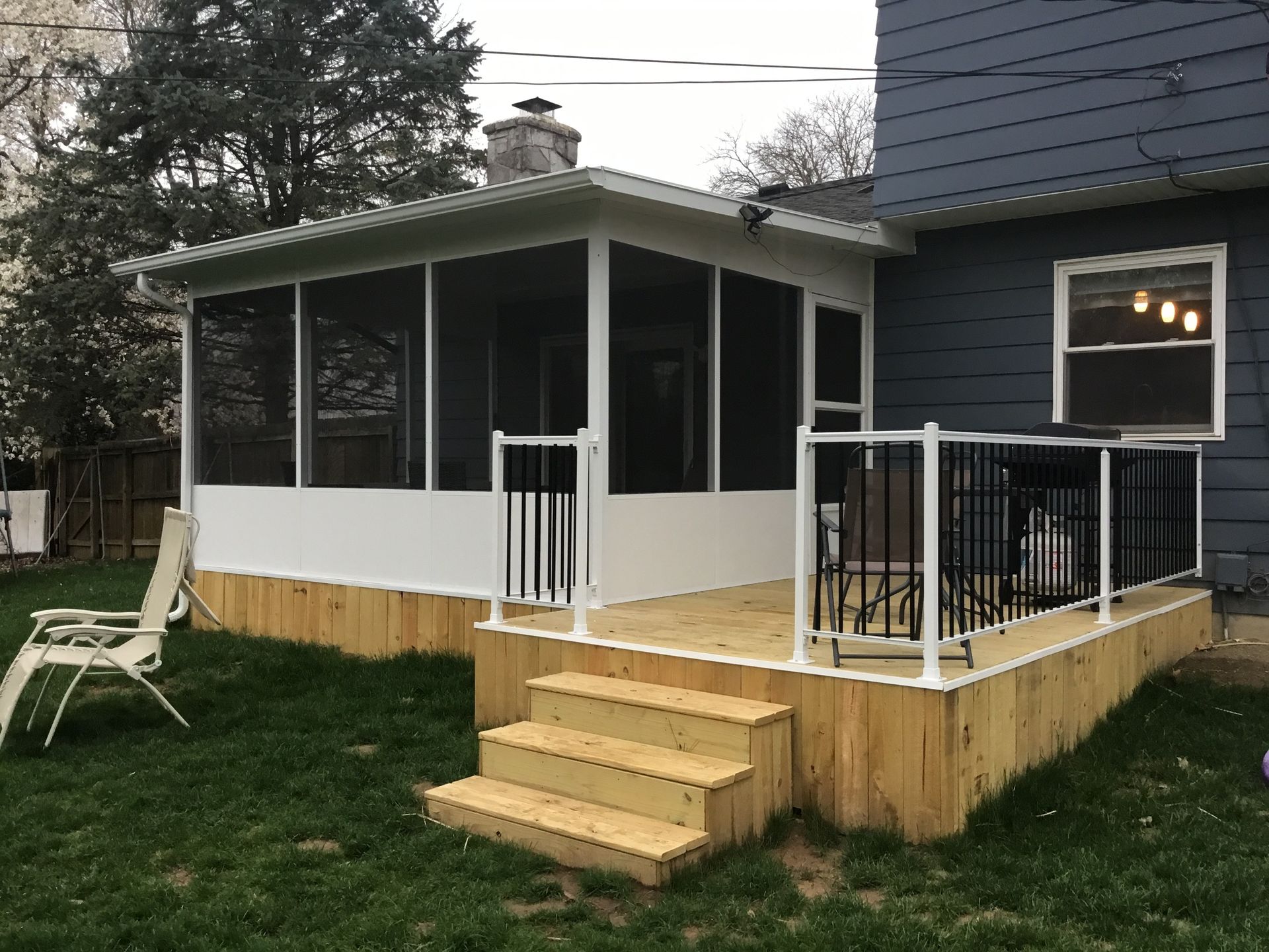 A newly built screened porch with a wooden deck and steps, attached to a gray house, set in a backyard.