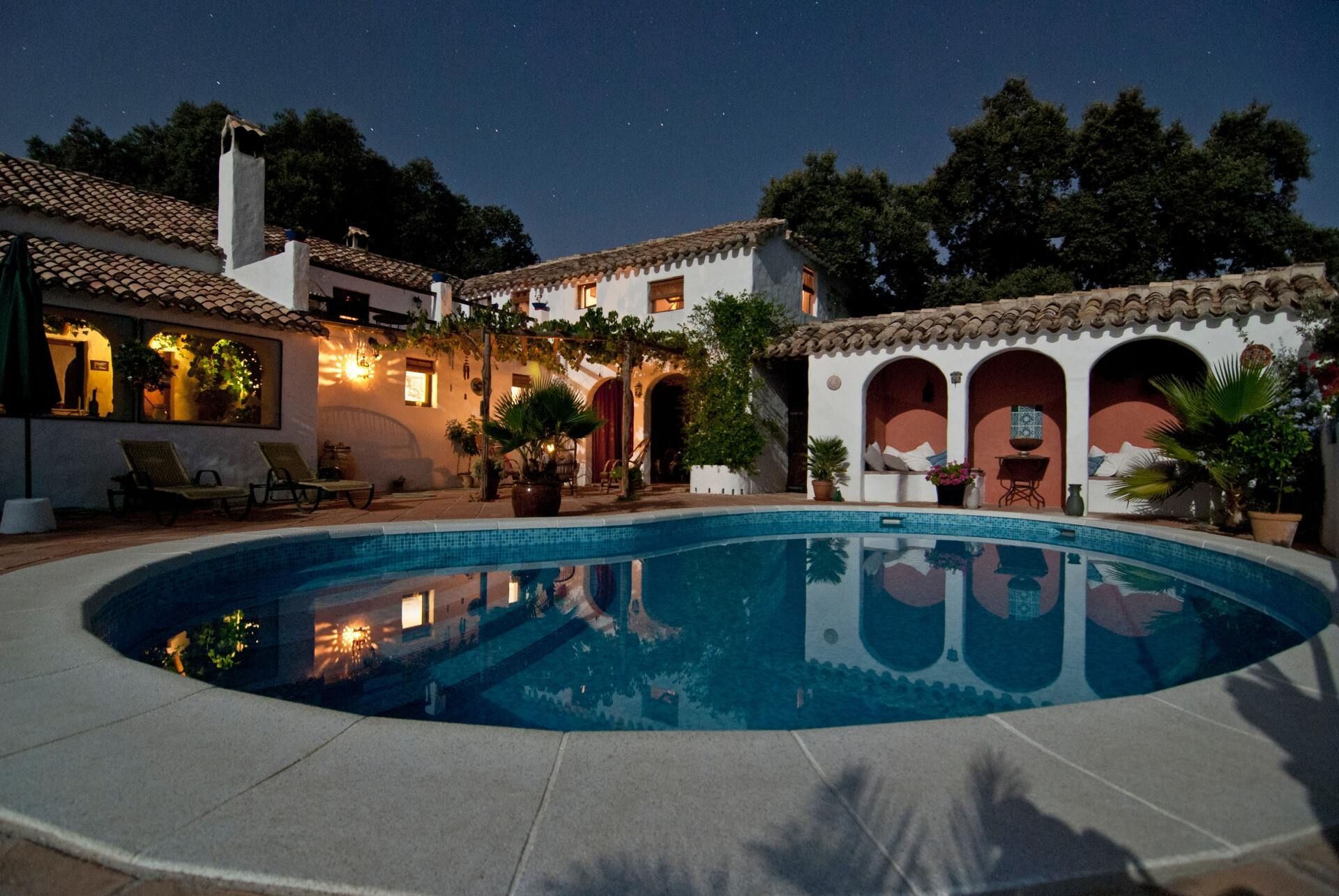 Nighttime view of a villa with a pool. White stucco buildings, arches, and a circular pool lit by interior and exterior lights.