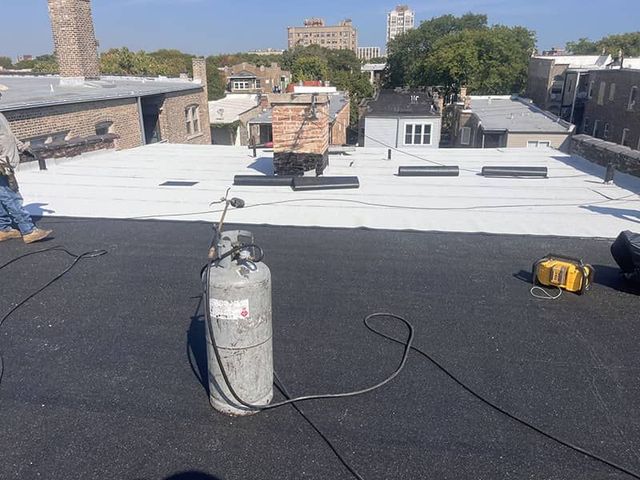 Flat rooftop with black-and-white roofing, a propane tank, and tools under a clear sky.