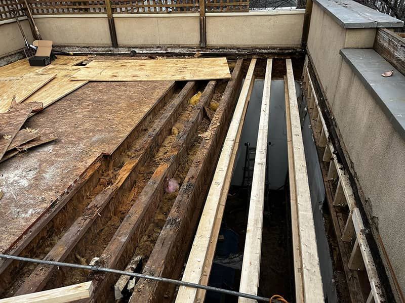 Rooftop construction site with exposed beams, plywood, and gravel beside a concrete wall.