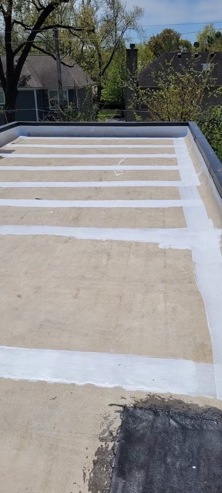 Flat rooftop with white membrane strips and tan patches, bordered by trees and a neighboring house.