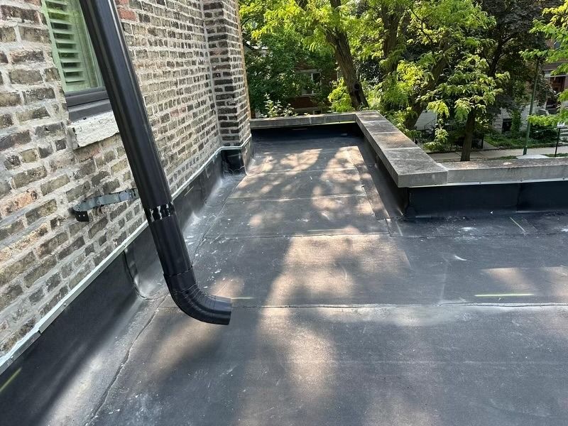 Rooftop patio with dark roofing, brick wall, downspout, and trees beyond the edge.