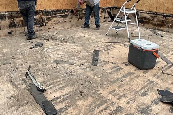 Workers removing flooring from a room with exposed subfloor, ladder, and tool box nearby