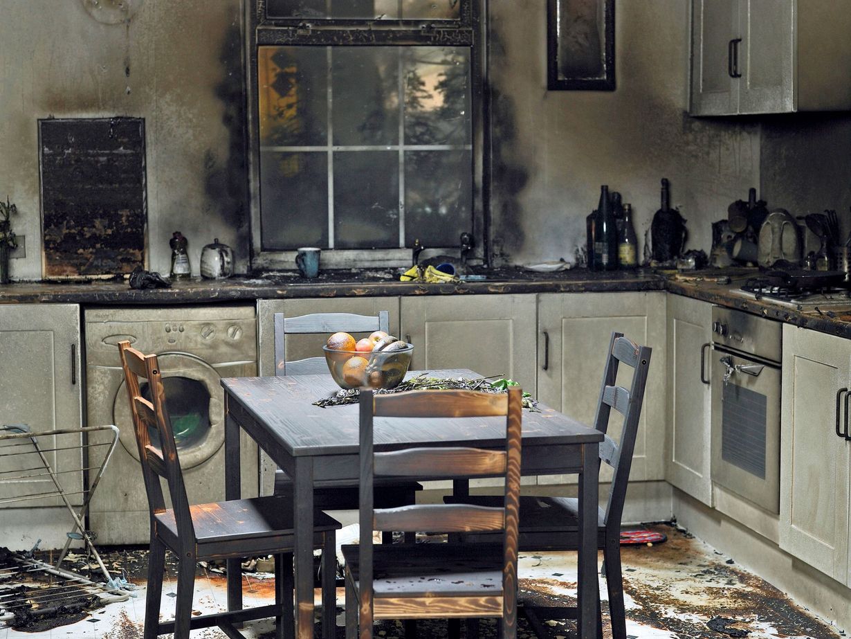 Kitchen interior destroyed by fire; charred cabinets, appliances, and table. Dark soot covers walls.