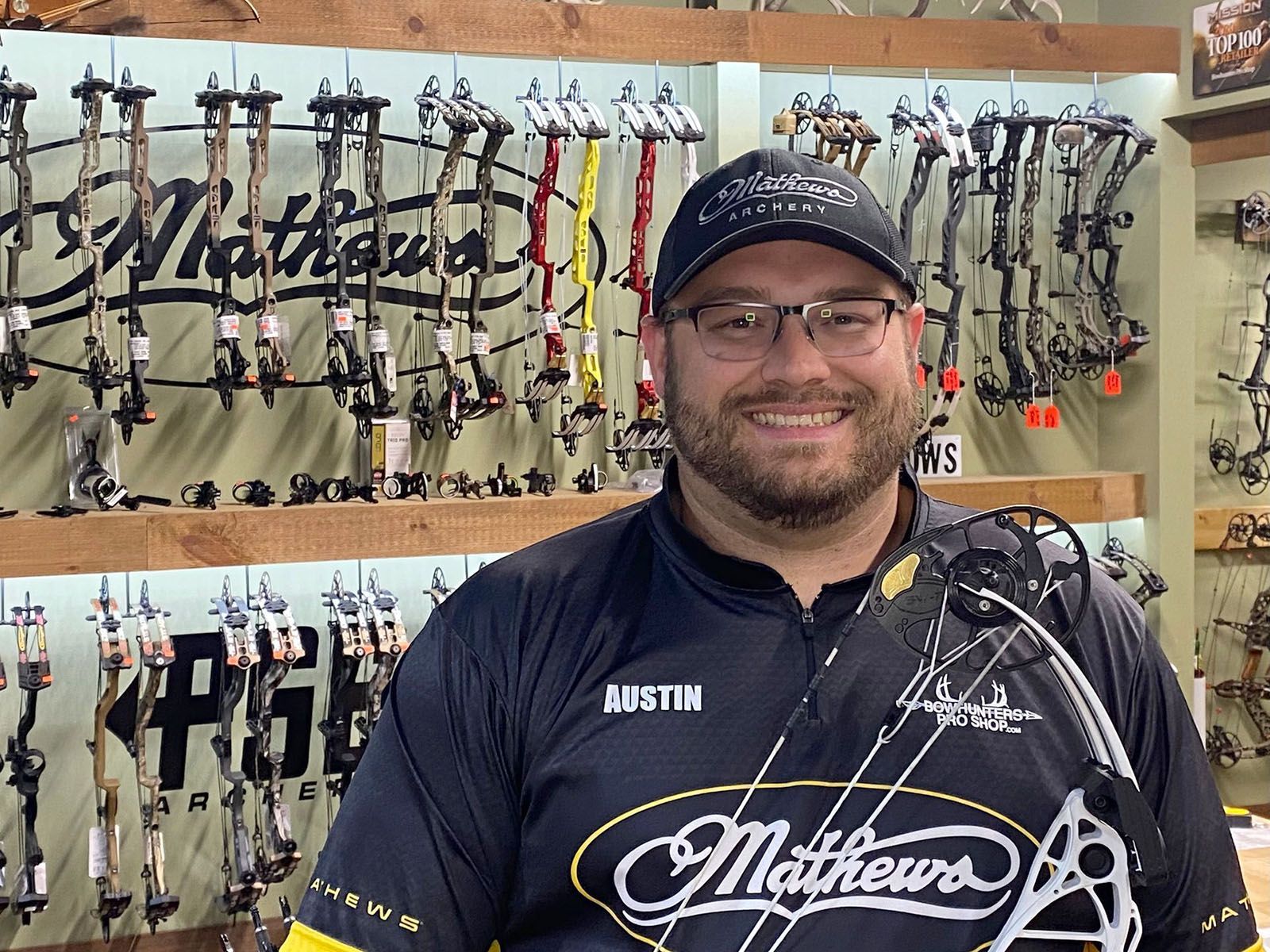 A man with eyeglasses in a black archery shirt holding a bow
