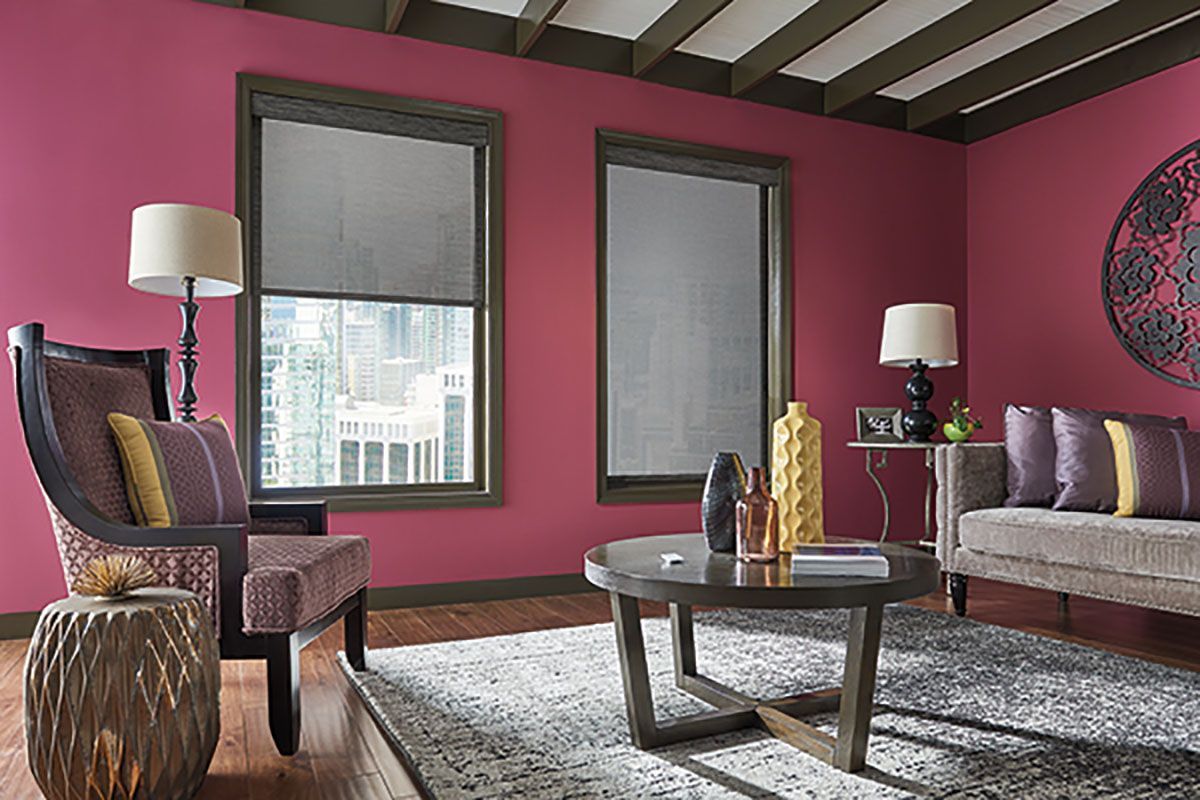 A living room with pink walls , a couch , a chair , a table and two windows.