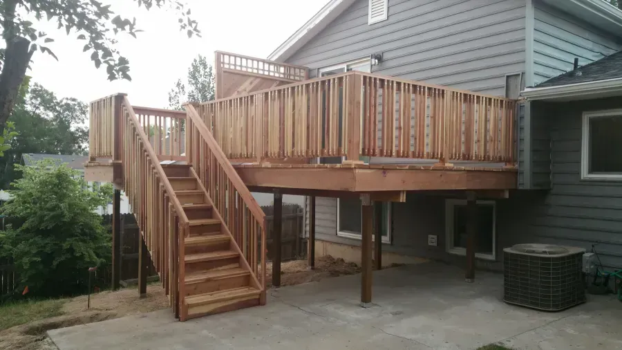 Wooden elevated deck with stairs beside a gray house in a backyard.
