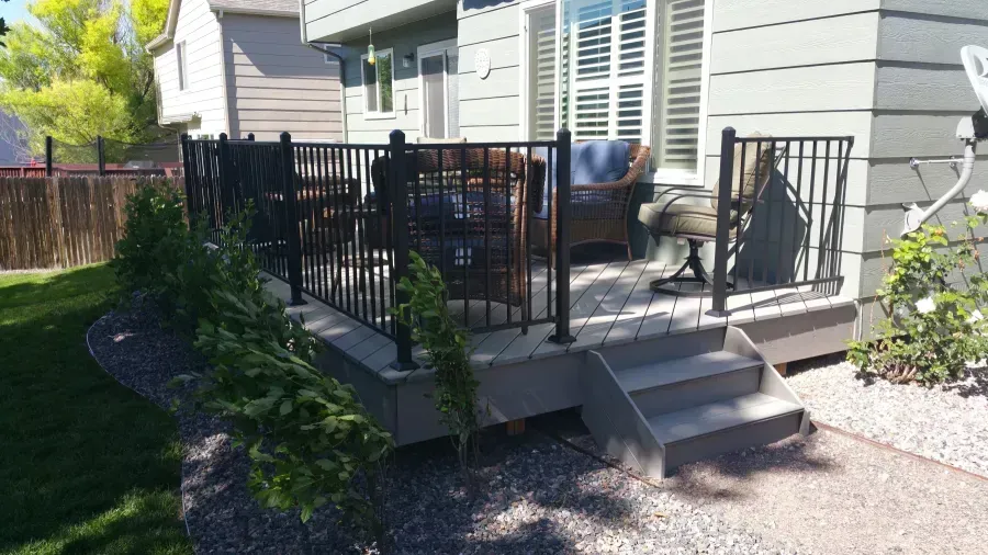 Patio deck with black railing, steps, grill, and chairs beside a gray house in a sunny yard