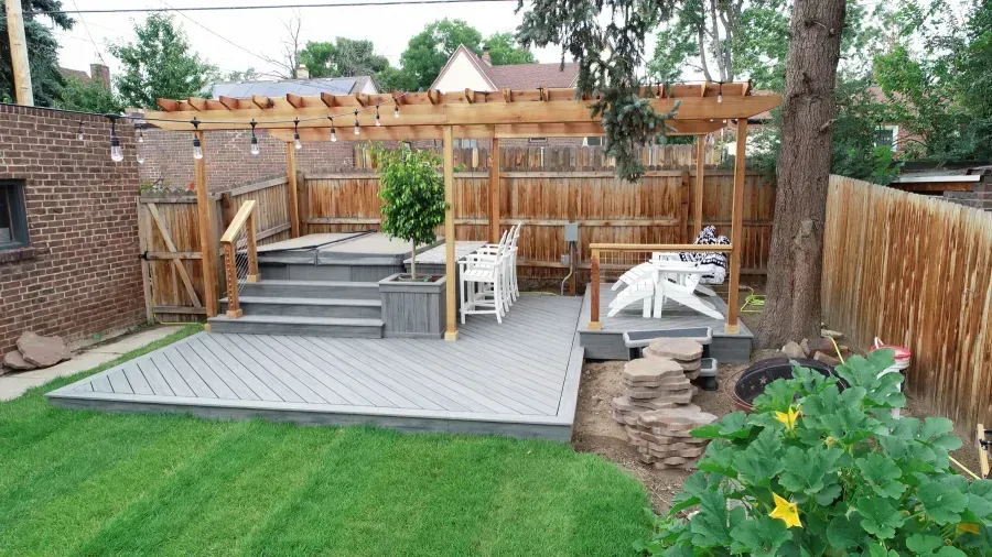 Backyard deck with gray seating area, wooden fence, steps, and green lawn with flowers
