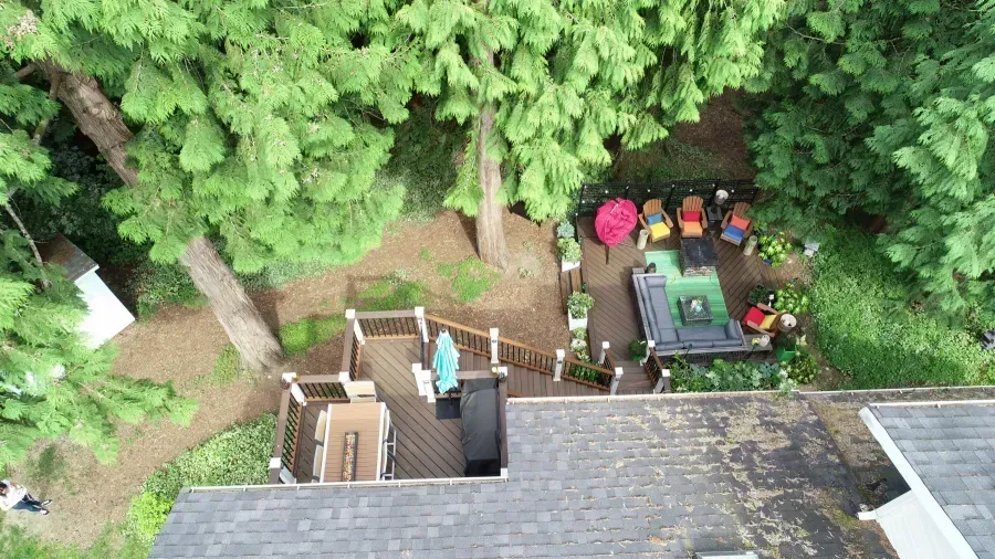 Overhead view of a backyard deck and patio under tall trees, with people gathered around a table and grill