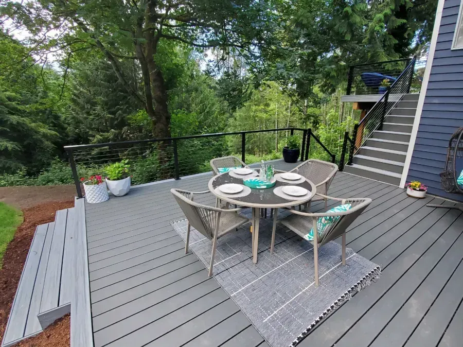 Outdoor deck with gray dining table and chairs, potted plants, and stairs beside a wooded yard.