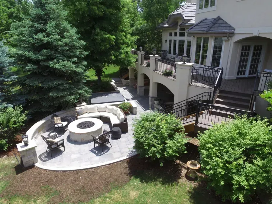 Backyard patio with circular fire pit seating beside a house and lush green trees