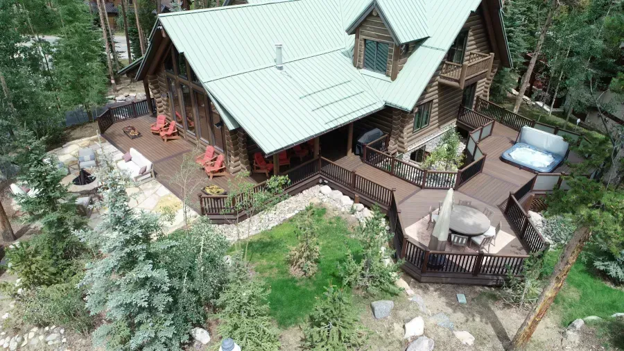 Aerial view of a rustic cabin with a green roof, wooden deck, and surrounding trees and garden.